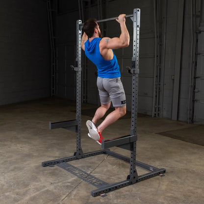 A person performing a pull-up on a strength training equipment in a gym setting, wearing a blue sleeveless hoodie and gray shorts.