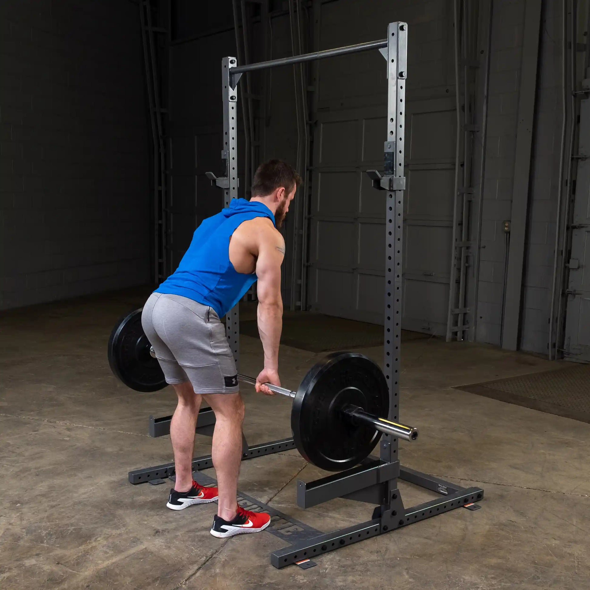 A man in a blue sleeveless hoodie and gray shorts is preparing to lift a barbell with weights in a gym setting, standing in front of a squat rack on a concrete floor.