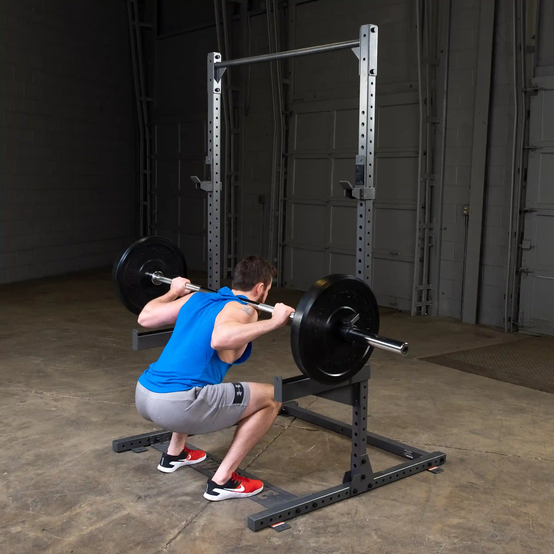 A man in a blue tank top and gray shorts performing a squat with a barbell on his shoulders in a gym setting.