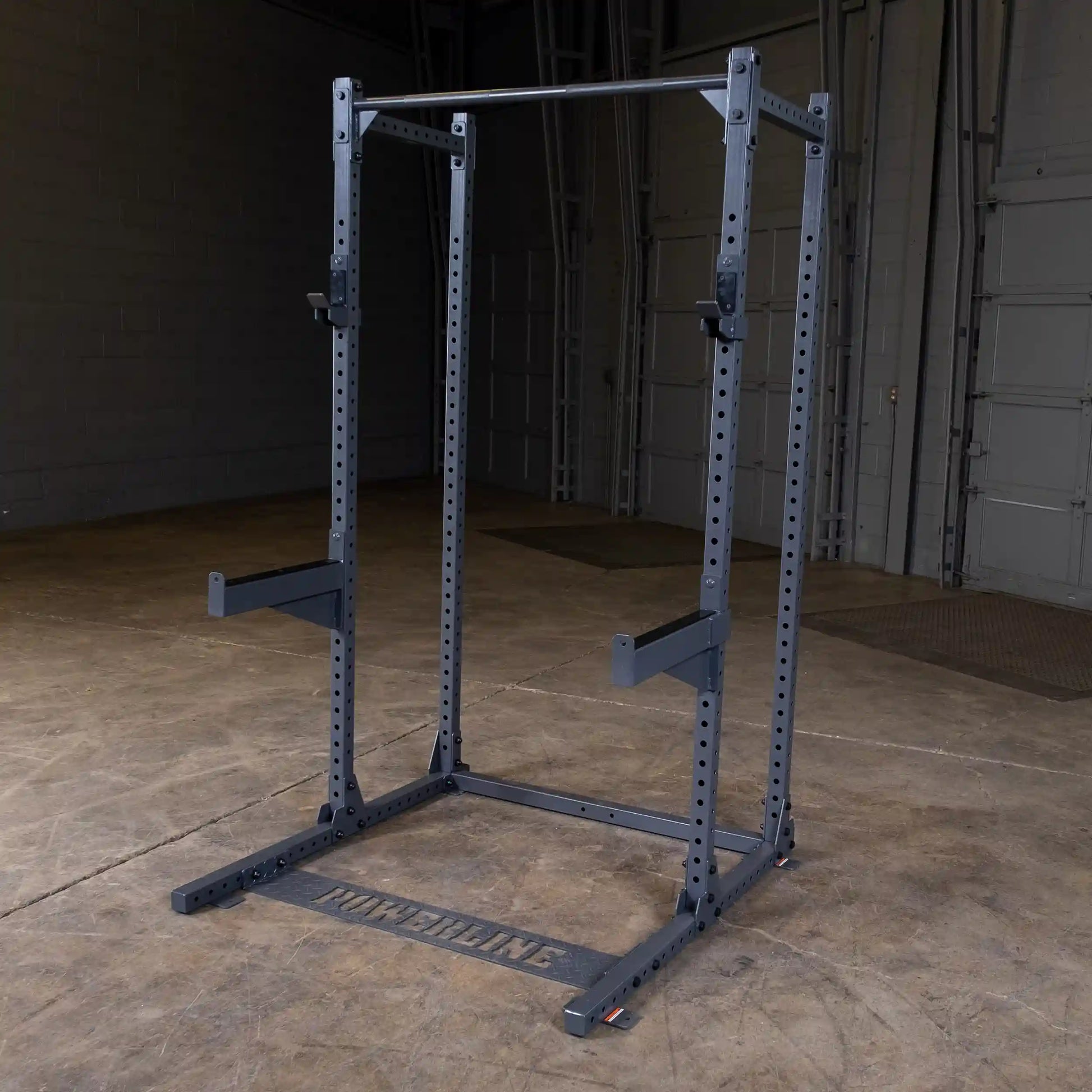 A power rack with a sturdy frame, featuring adjustable barbell holders and a pull-up bar, set in an empty gym space with a concrete floor and gray walls.