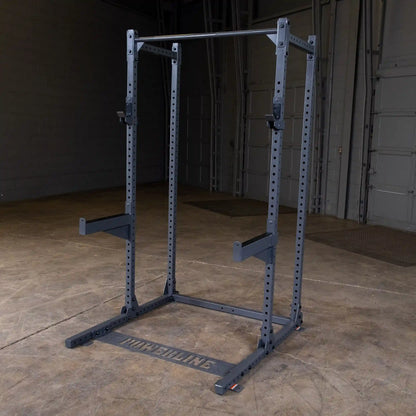 A power rack with a sturdy frame, featuring adjustable barbell holders and a pull-up bar, set in an empty gym space with a concrete floor and gray walls.