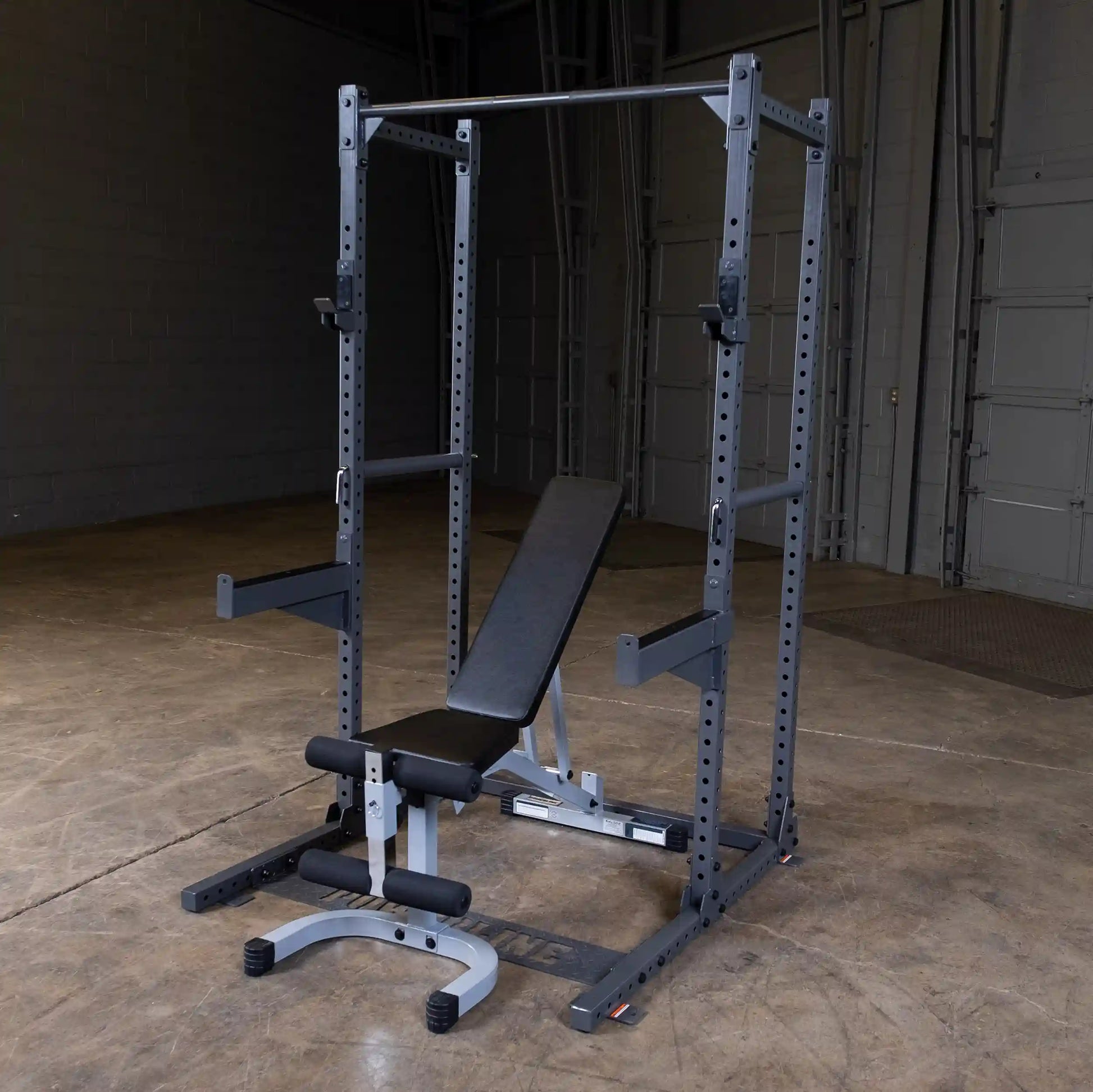 A black and silver weight bench and squat rack set up in an empty gym space with concrete flooring and gray walls.