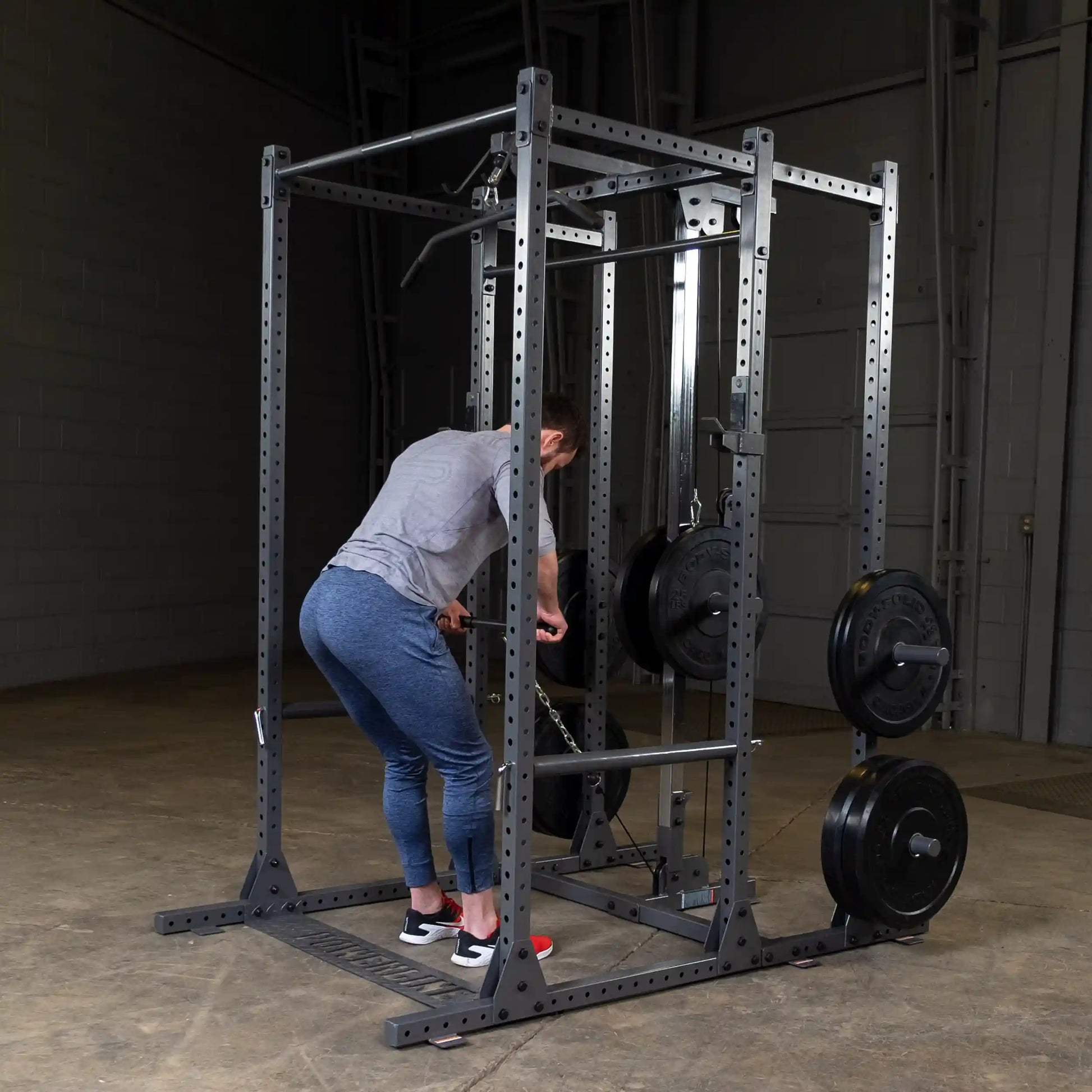A person in a gray shirt and blue leggings is performing an exercise using a weightlifting cage, adjusting weights on the bar. The gym setting has a concrete floor and metal equipment.