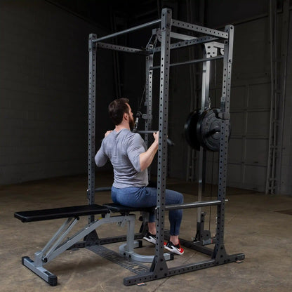 A man in a gray long-sleeve shirt and blue pants performs a seated exercise on a cable machine in a gym setting, with a weight plate visible on the machine.