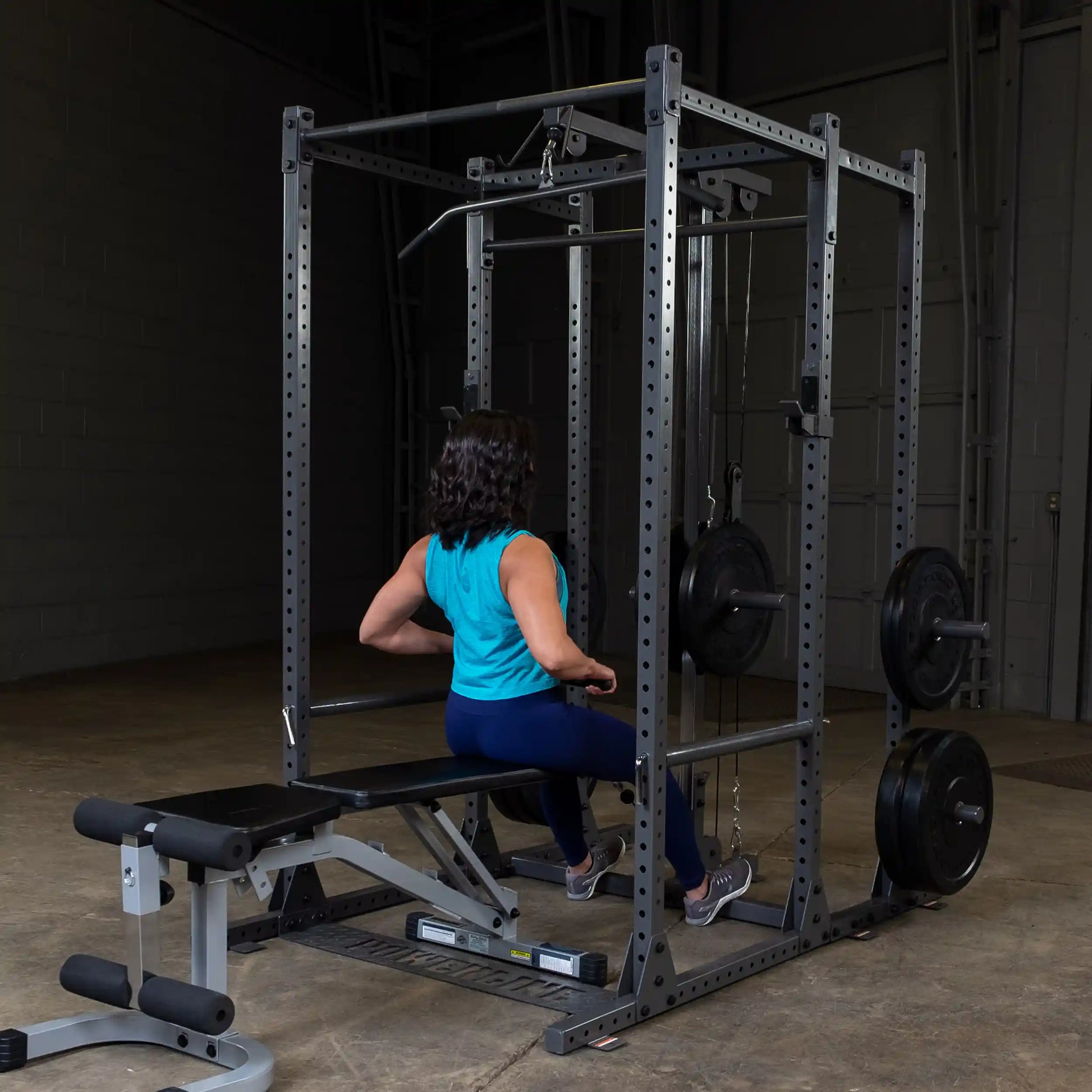 A person in a blue tank top and leggings is seated on a weight bench, using a cable machine in a gym setting. The gym features a squat rack with weights and a dimly lit background.