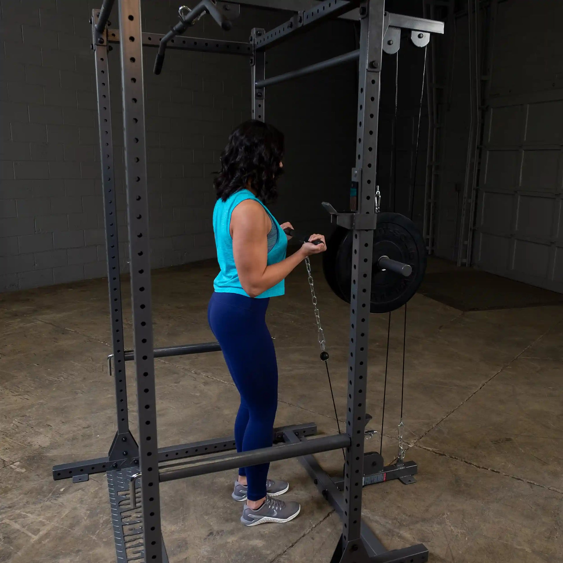 A person in a turquoise tank top and blue leggings is using a cable machine in a gym, preparing for a workout.
