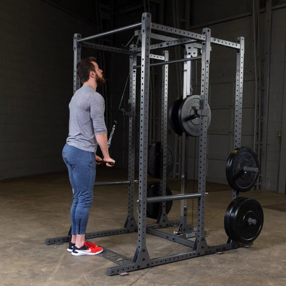 A man in a gray long-sleeve shirt and blue leggings performs a cable exercise in a gym, using a cable machine with weights attached. He is focused on his workout, standing on a concrete floor with gym equipment in the background.
