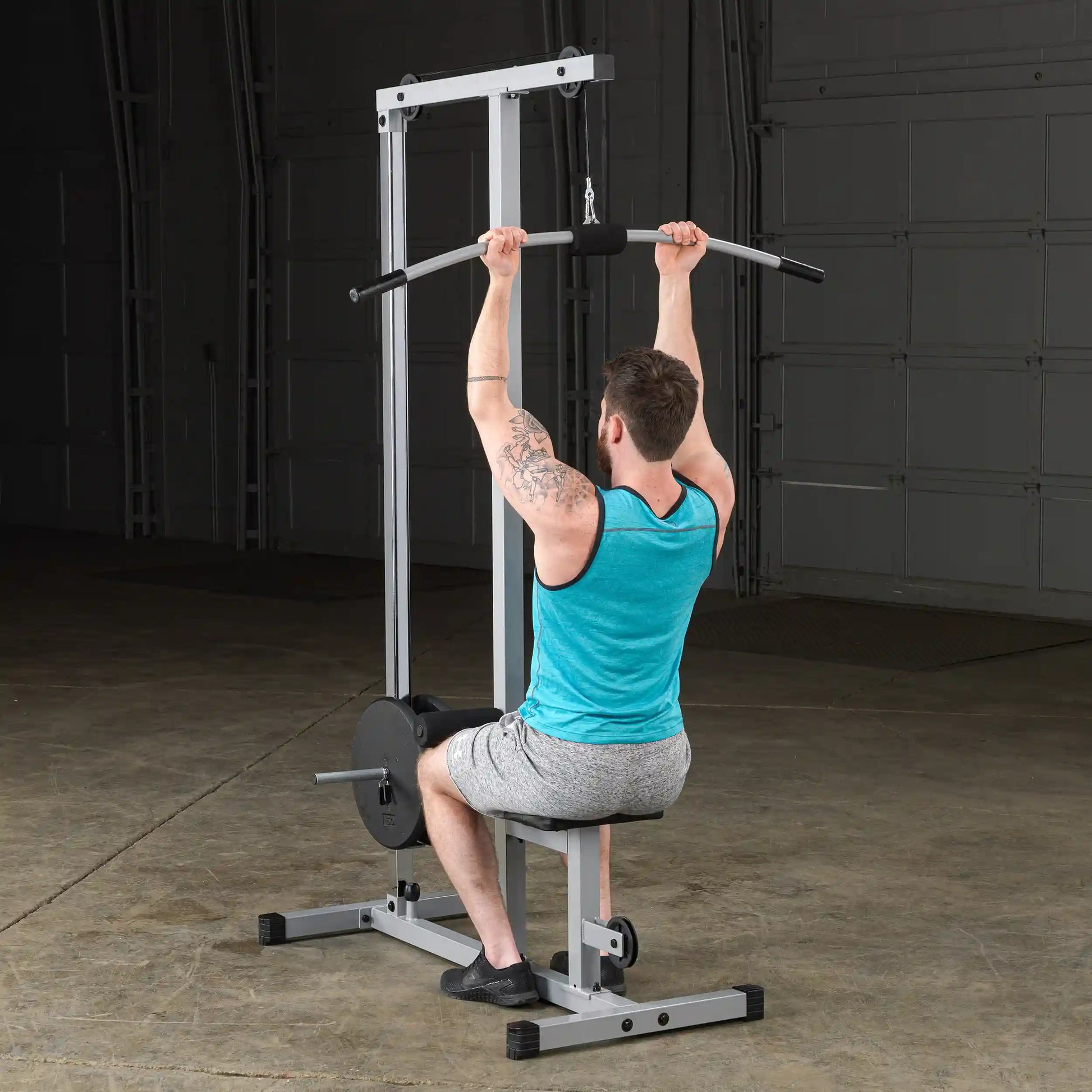 A man with tattoos performs a seated pull-down exercise on a gym machine, wearing a blue tank top and gray shorts, in a spacious indoor gym setting.