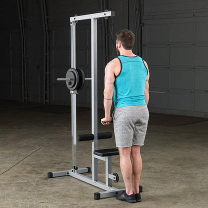 A man in a turquoise tank top and gray shorts stands in front of a weightlifting machine, preparing to perform an exercise. The setting is a gym with a concrete floor and a neutral background.