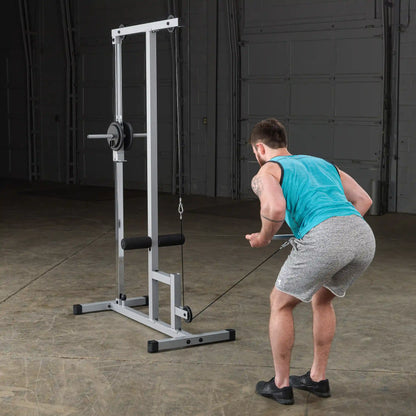 A person in a blue tank top and gray shorts performs a cable row exercise using a cable machine in a gym setting. The background is dimly lit, highlighting the equipment and the individual's form.