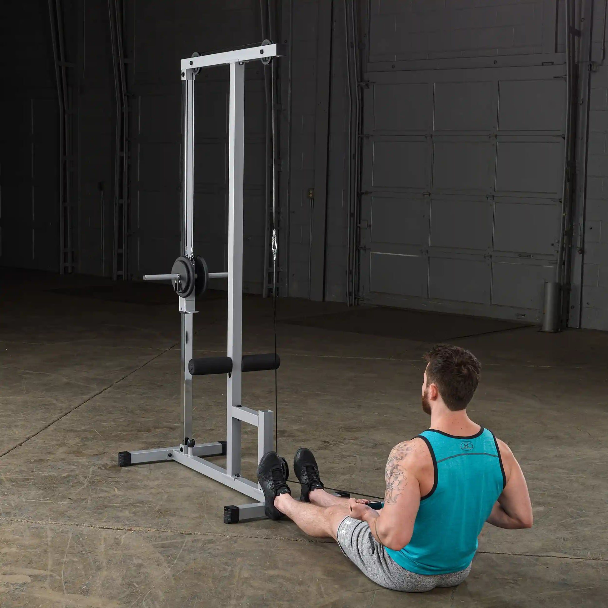A man sitting on the floor in a gym, performing a seated row exercise using a cable machine. The machine has a vertical frame with a weight plate and a cable attached. The man is wearing a teal tank top and gray shorts, with a focus on his form and the equipment in a spacious, well-lit gym environment.