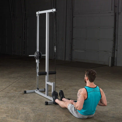 A man sitting on the floor in a gym, performing a seated row exercise using a cable machine. The machine has a vertical frame with a weight plate and a cable attached. The man is wearing a teal tank top and gray shorts, with a focus on his form and the equipment in a spacious, well-lit gym environment.