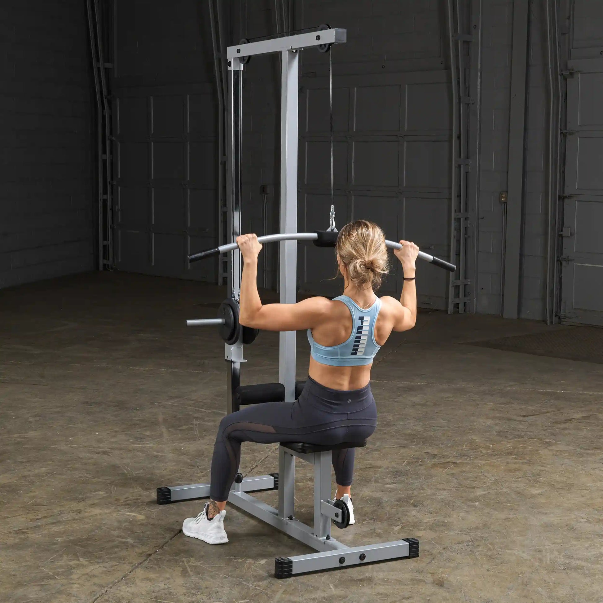 A woman with a ponytail is seated on a weight machine, performing a lat pulldown exercise. She is wearing a light blue sports bra and black leggings, focusing on her workout in a spacious gym environment.
