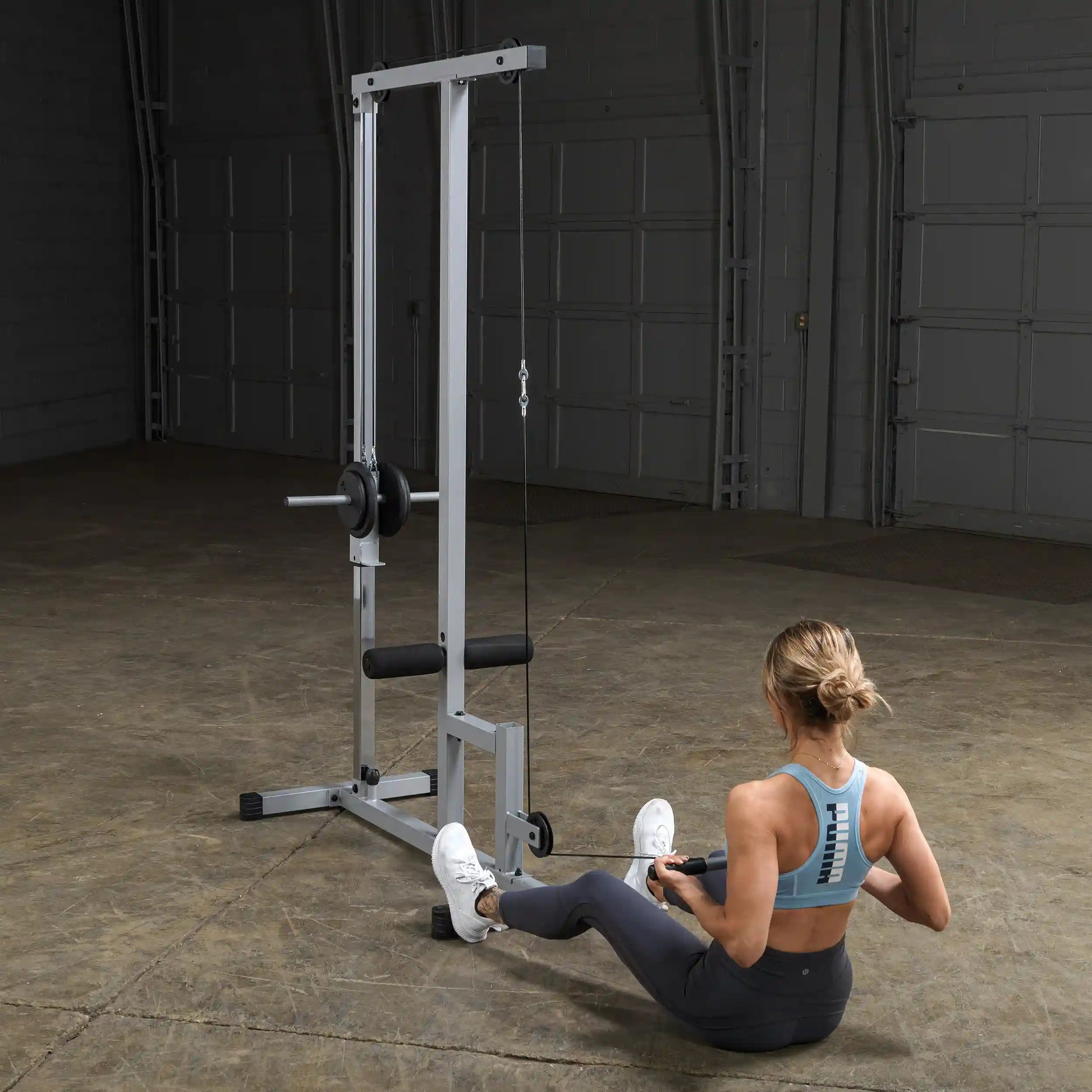 A woman exercising on a cable machine in a gym setting, performing a seated row. She is wearing a light blue sports bra and dark leggings, with her hair styled in a loose bun.