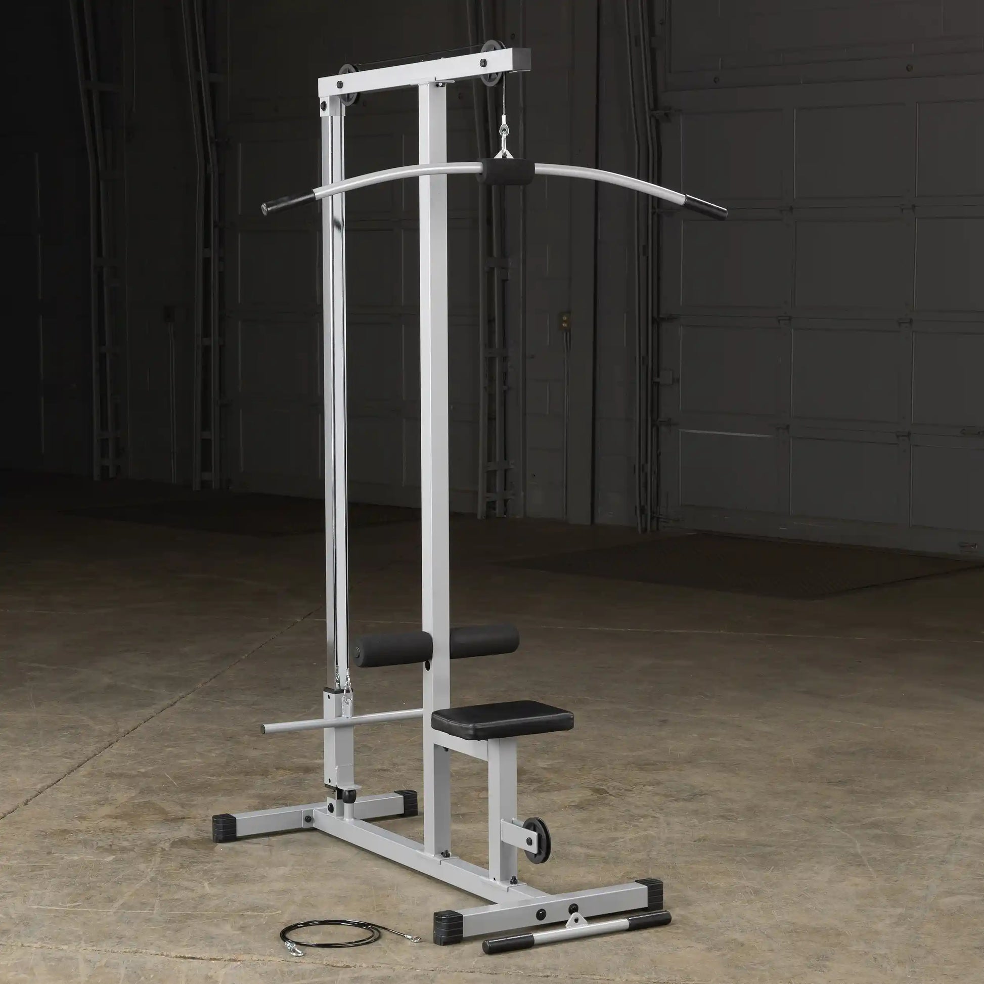 A silver cable machine for strength training, featuring a curved bar attachment, a seat, and foam rollers, set against a dark background in a gym or workout space.