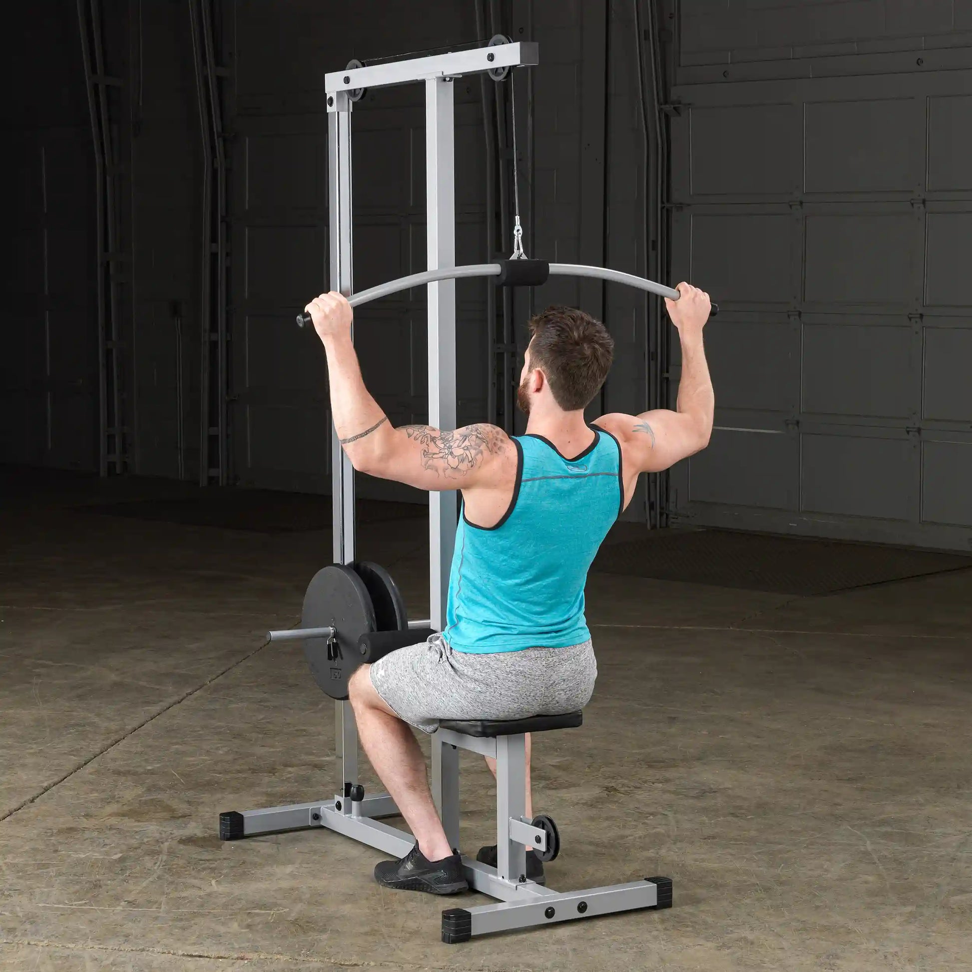 A man with a tattoo is performing a lat pulldown exercise on a fitness machine in a gym setting, wearing a turquoise tank top and gray shorts.