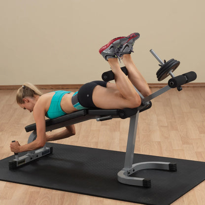 A woman performing a leg exercise on a workout bench, positioned on her stomach with her legs raised and bent, wearing a turquoise sports top and black shorts, in a gym setting.