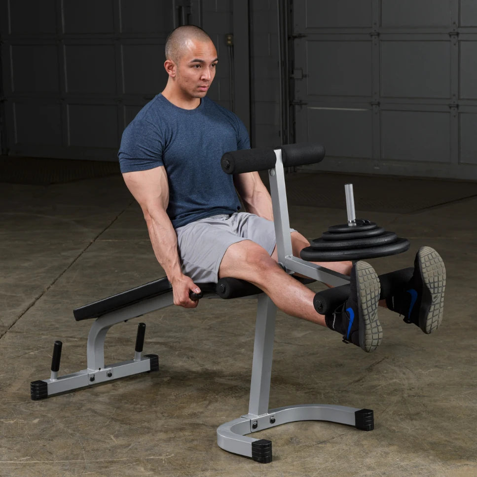 A man performing a leg exercise on a weight bench in a gym, using a leg extension machine with weights, wearing a blue t-shirt and gray shorts.