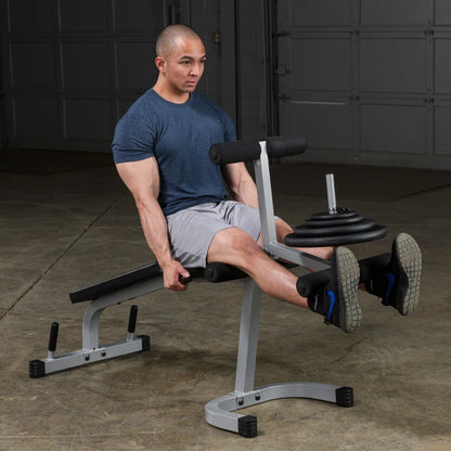 A man performing a leg exercise on a weight bench in a gym, using a leg extension machine with weights, wearing a blue t-shirt and gray shorts.