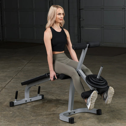 A woman with long blonde hair sits on a weight bench, performing an exercise. She wears a black crop top and olive green leggings, gripping the handles of the machine with weights attached. The background is a gray garage setting.