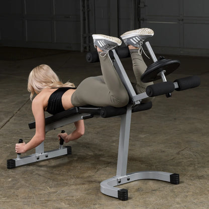 A person performing an exercise on a gym equipment designed for targeting the lower back and hamstrings, positioned face down with legs elevated, in a fitness studio setting.