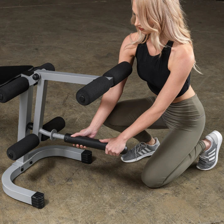 A woman in athletic wear kneeling next to a gym equipment, adjusting a barbell attachment. She has long blonde hair and is focused on her task.