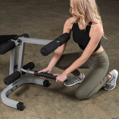 A woman in athletic wear kneeling next to a gym equipment, adjusting a barbell attachment. She has long blonde hair and is focused on her task.