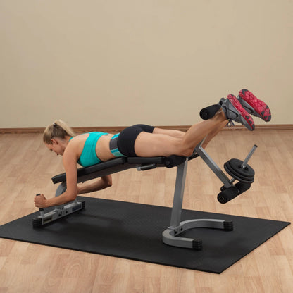 A woman in a turquoise sports bra and black shorts performs a leg exercise on a workout bench, focusing on her hamstrings, with weights attached to the bench in a fitness studio.