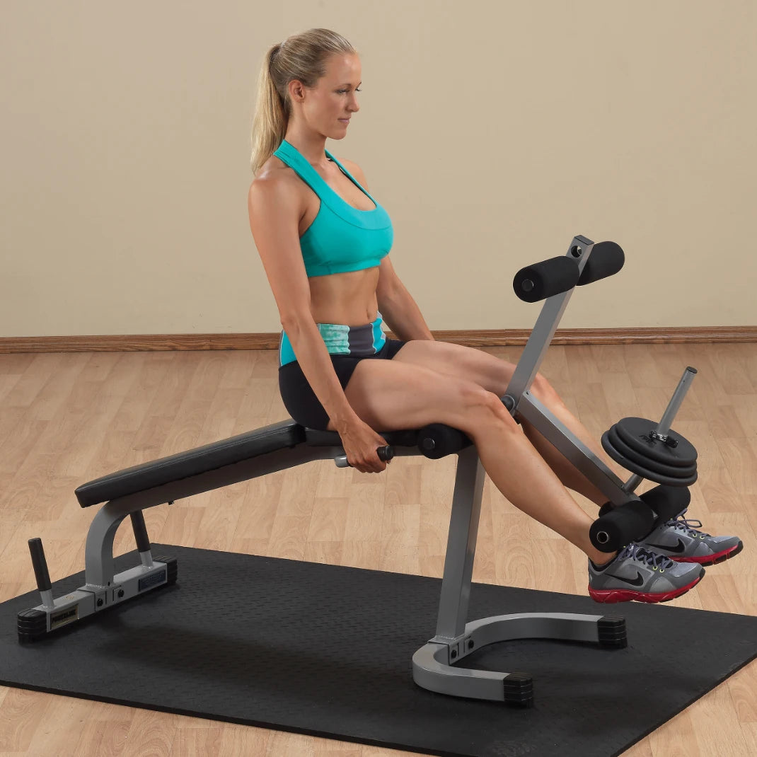 A woman in a turquoise sports bra and black shorts is performing an exercise on a weight bench, targeting her legs. The setup includes weights on one side of the bench, with a rubber mat on the floor.