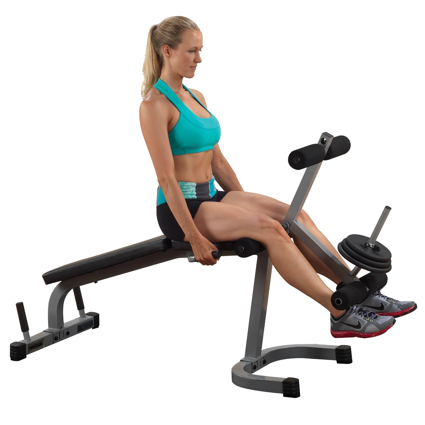 A woman in a turquoise sports bra and black shorts is performing a seated leg curl exercise on a workout bench, demonstrating proper form while gripping the handles.