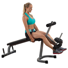 A woman in a turquoise sports bra and black shorts is performing a seated leg curl exercise on a workout bench, demonstrating proper form while gripping the handles.