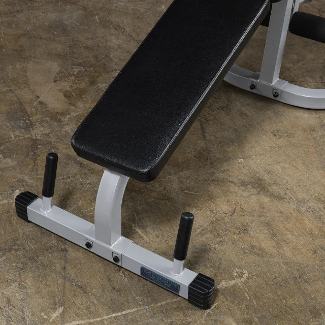 A close-up view of a black padded weight bench with a sturdy silver frame, featuring adjustable height pegs on either side, set against a textured floor surface.
