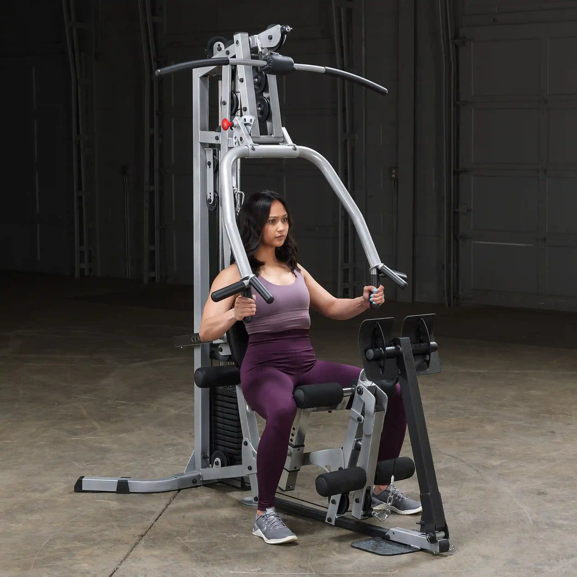 A woman wearing a purple tank top and leggings is seated on a multi-gym machine, preparing to perform an exercise, in a spacious indoor setting.