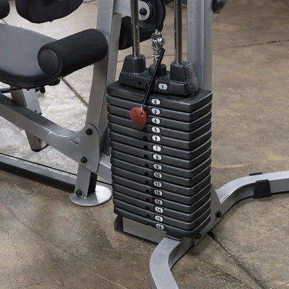A close-up of a weight stack on a multi-gym exercise machine, showing numbered weight plates from 1 to 16 and a red selector pin. The machine has a gray and black color scheme, with a padded seat visible in the background.