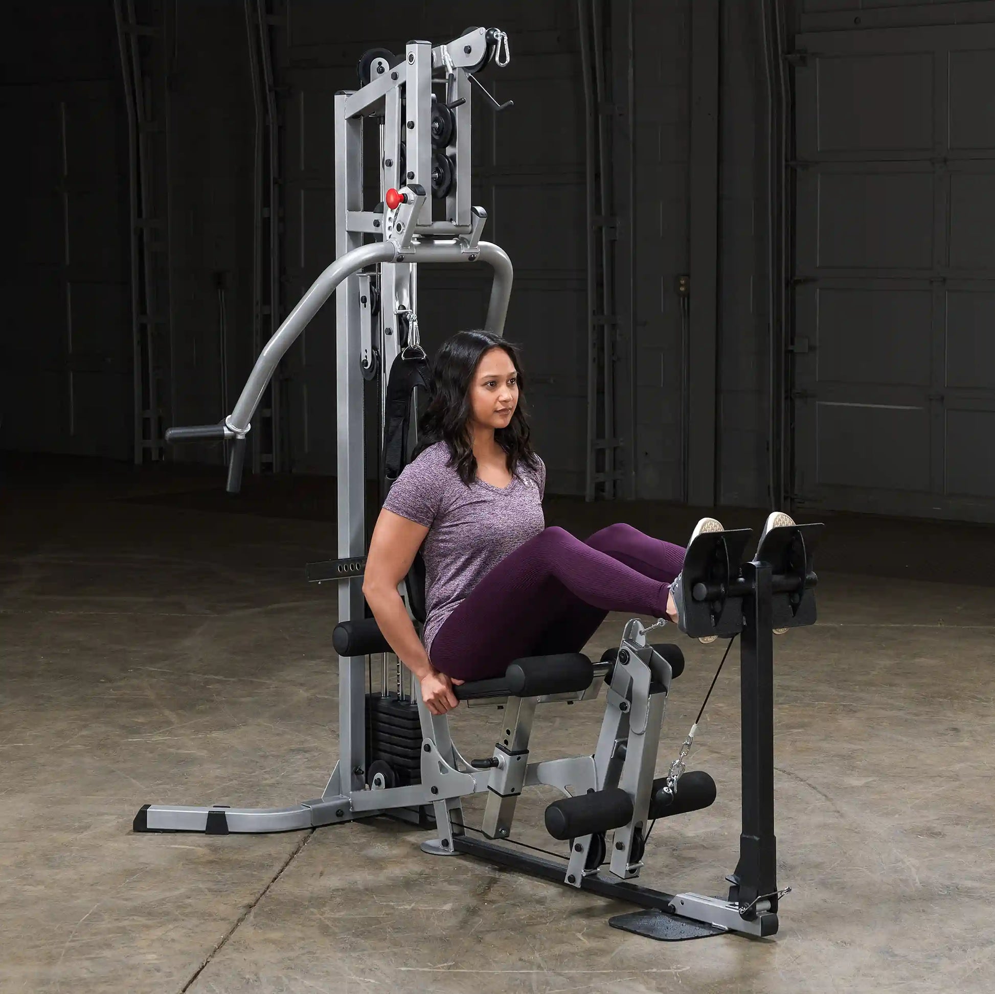 A woman exercising on a multi-gym machine, performing leg curls. She is wearing a purple shirt and leggings, with a focused expression, in a spacious gym setting.
