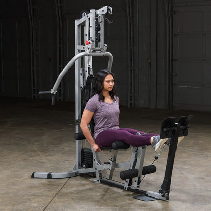 A woman performing leg exercises on a multi-gym machine in a spacious indoor gym setting. She is seated with her legs extended, wearing a purple shirt and leggings, focusing on her workout.