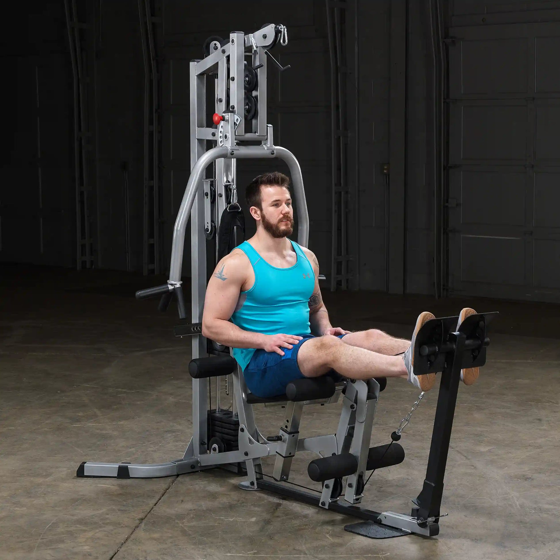 A man in a turquoise tank top and shorts is seated on a gym machine, preparing to perform a leg exercise. The machine is set up in a spacious indoor area with a neutral background.