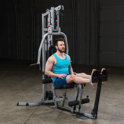 A man in a turquoise tank top and shorts is seated on a gym machine, preparing to perform a leg exercise. The machine is set up in a spacious indoor area with a neutral background.