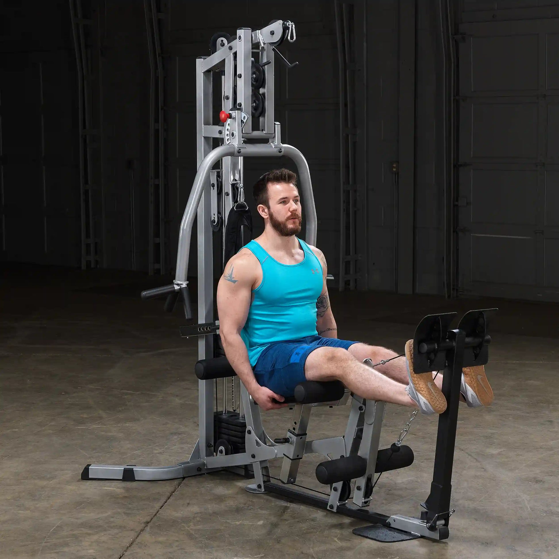 A man in a turquoise tank top and blue shorts performs leg exercises on a weight machine in a gym setting.