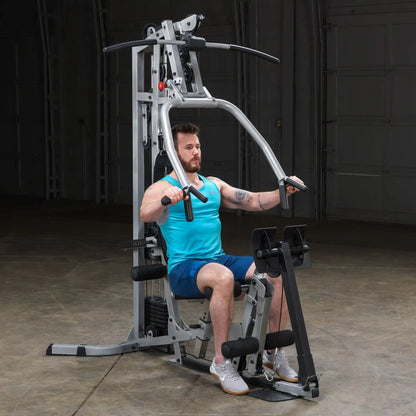 A man with a beard is seated on a gym machine, wearing a blue tank top and shorts, preparing to perform an exercise. The machine is set up for strength training, with various handles and attachments visible in a spacious, dimly lit gym environment.