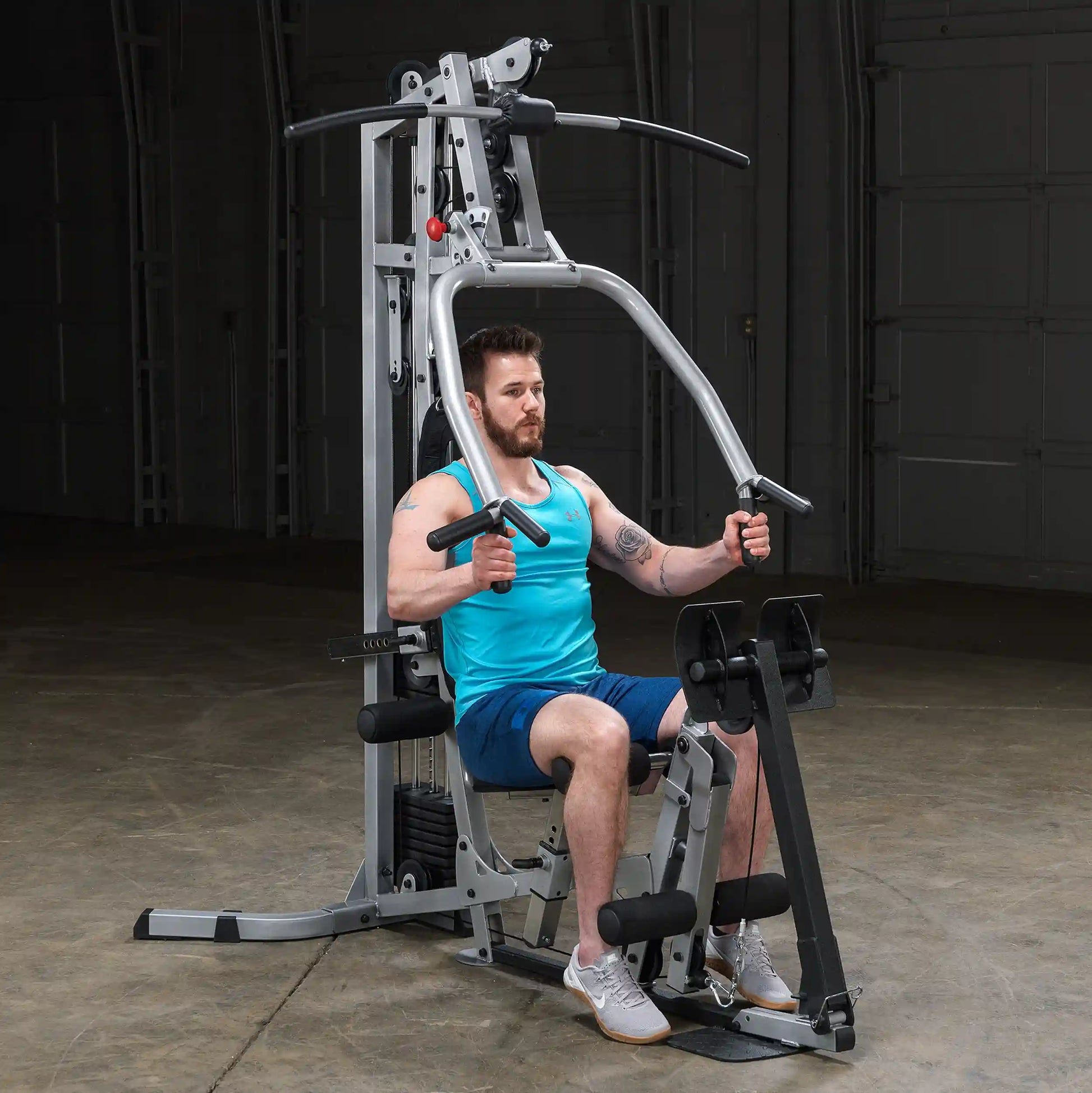 A man with a beard wearing a turquoise tank top and blue shorts is exercising on a multi-gym machine in a dimly lit room.