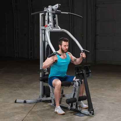 A man in a turquoise tank top and shorts exercises on a multi-gym machine, focusing on strength training. The gym is indoors with a concrete floor and a simple background.