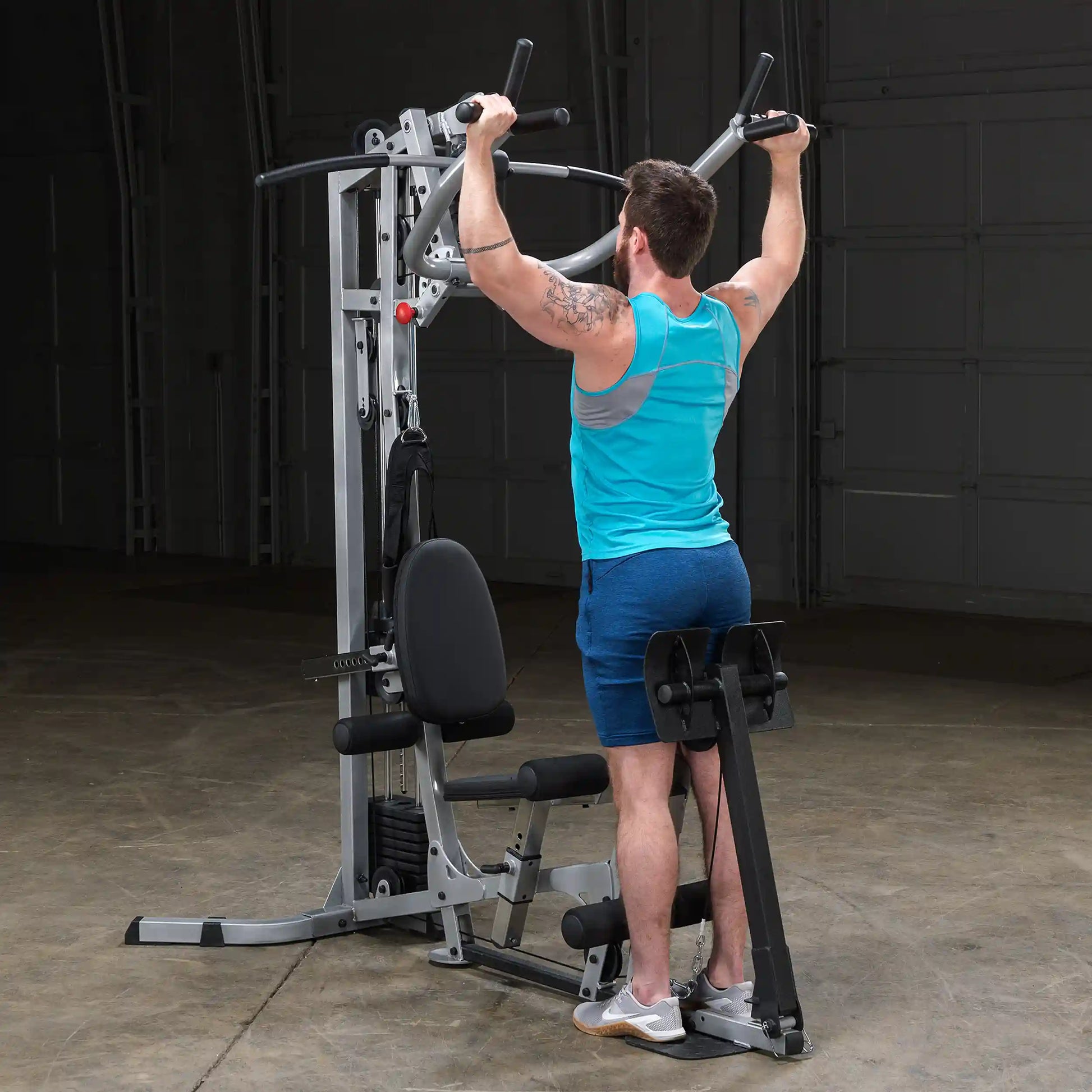 A man wearing a blue tank top and shorts performs a pull-up on a multi-gym machine in a spacious gym setting.