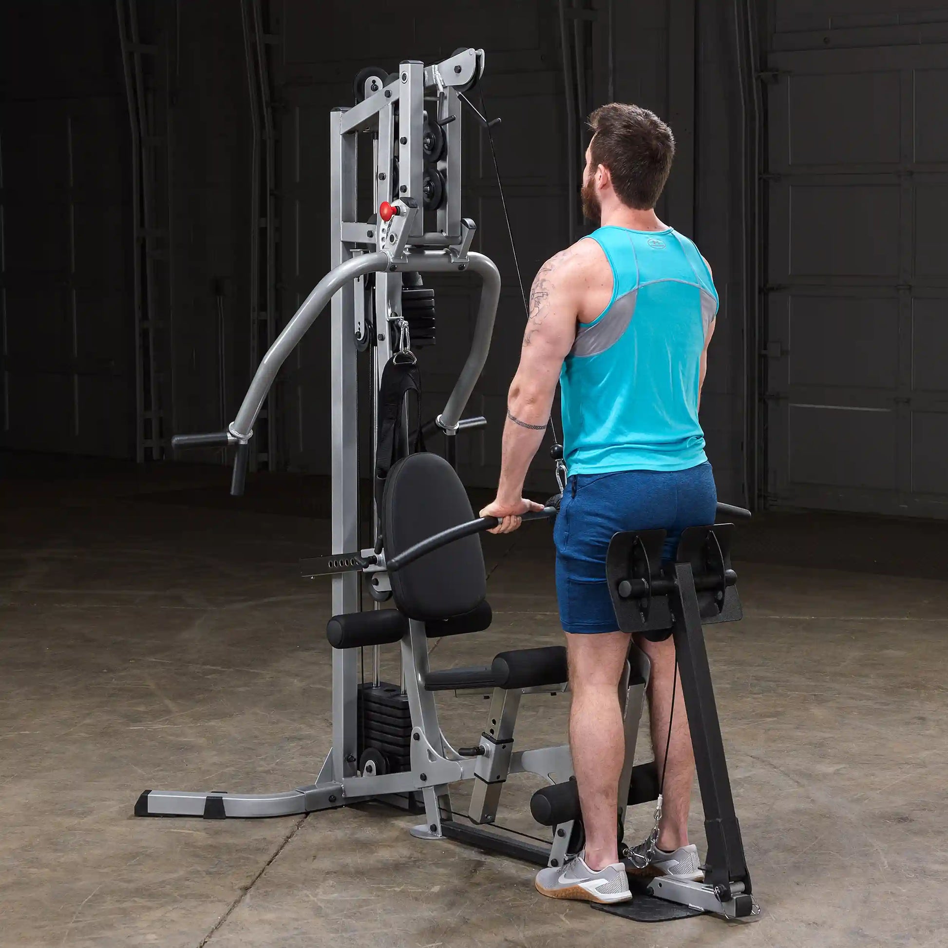 A man in a blue tank top and shorts using a multi-gym exercise machine in a dimly lit gym setting.