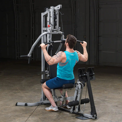A man with a tattoo, wearing a blue tank top and shorts, exercises on a multi-gym machine, performing a lat pull-down in a well-lit gym environment.