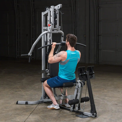 A man with a beard and tattoos is using a multi-gym machine for a workout, sitting on a bench and pulling a bar towards him in a dimly lit gym space.