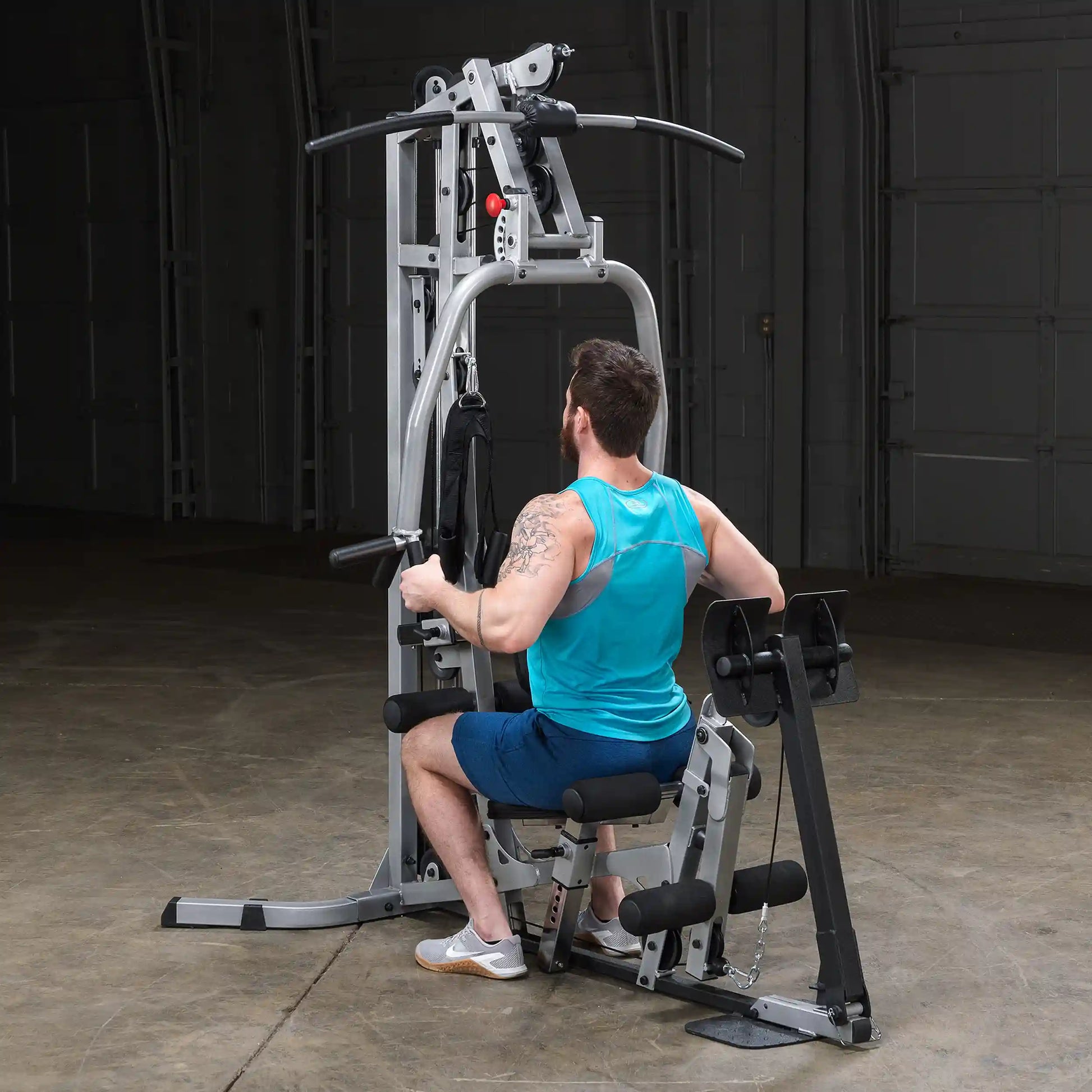 A man with a beard wearing a blue tank top and shorts is seated on a multi-gym machine, performing an exercise. The gym environment is well-lit with a neutral background.