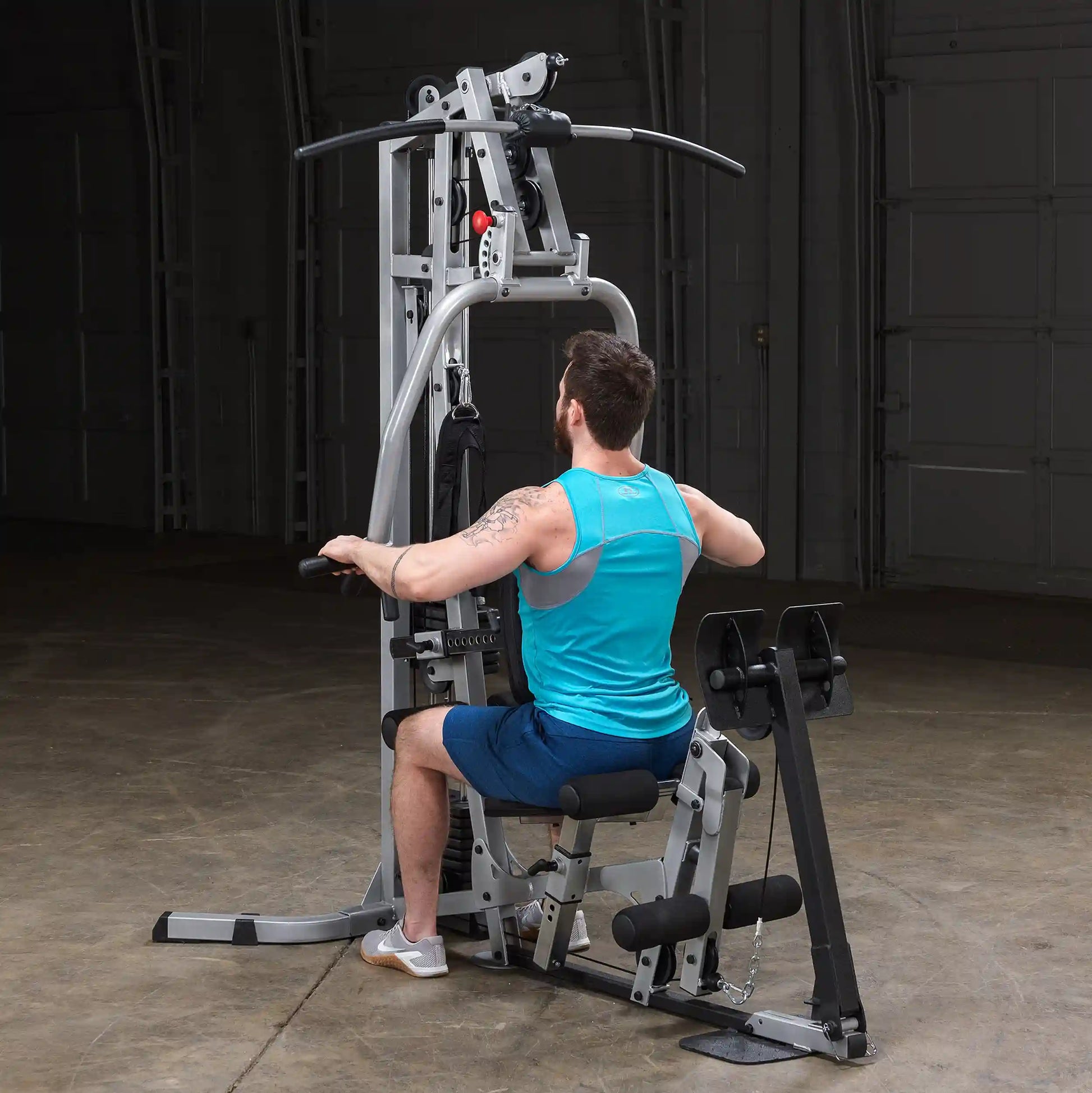 A man with a beard wearing a blue tank top and shorts is seated on a multi-gym machine, performing an exercise. The gym equipment features various pulleys and handles, set in a spacious, industrial-style gym environment.