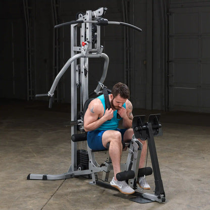 A man with a beard is seated on a multifunctional gym machine, preparing to perform an exercise. He is wearing a blue tank top and shorts, focusing on his form as he grips the handles. The gym setting has a neutral background with ample space around the equipment.