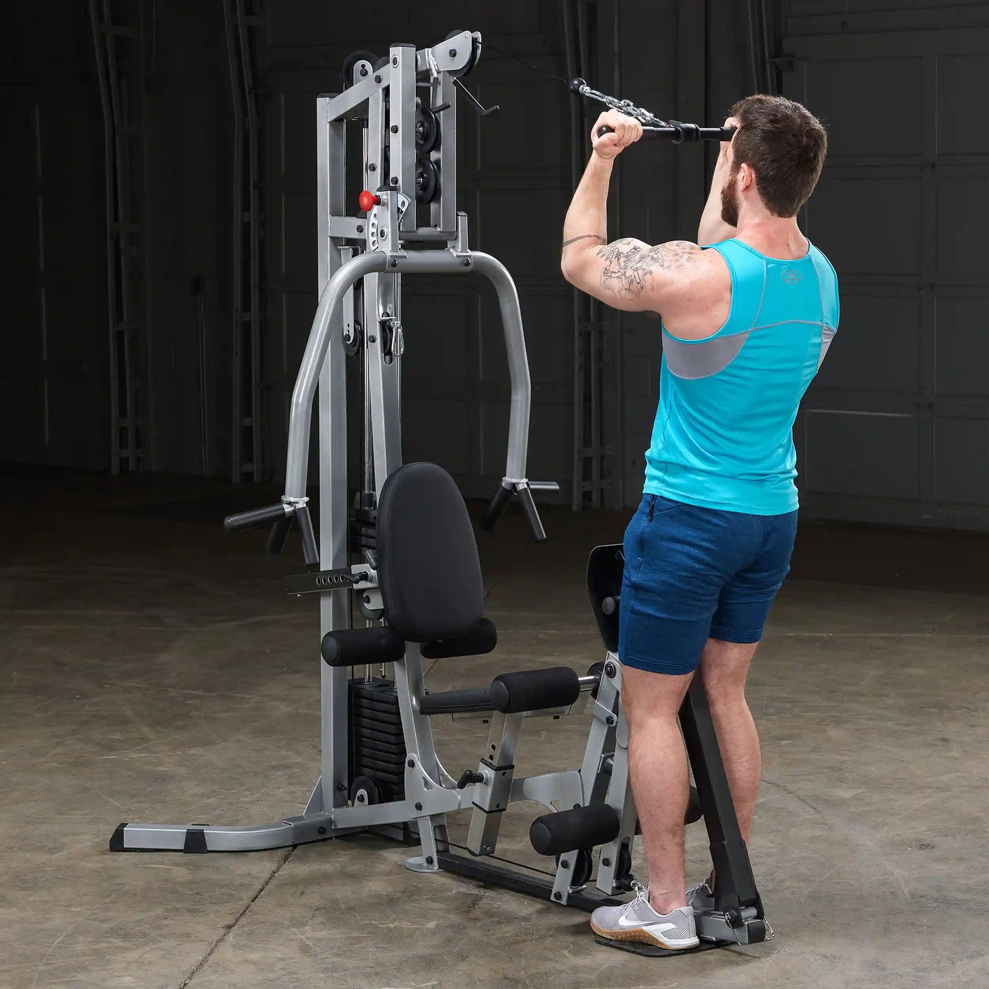 A man in a turquoise tank top and blue shorts performs a lat pull-down exercise on a multi-gym machine in a spacious indoor gym setting.